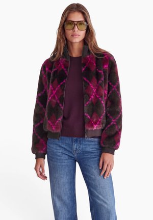 Femme portant des lunettes de soleil teintées jaunes, un pull violet foncé, un jean bleu et une veste en fourrure foncée avec un motif losange rose.