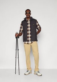 Pilka be rankovių liemenė dėvima ant languotosios marškinių, smėlio spalvos kelnės ir šviesūs sportiniai bateliai. Laiko juodus žygių lazdas. Smėlio spalvos saulės akiniai papildo įvaizdį.