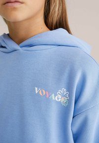 Sweat à capuche bleu clair avec une texture douce, présentant un design de texte "VOYAGE" multicolore et des accents floraux sur la zone gauche de la poitrine.