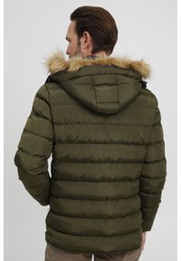 Oliven grønn pufferjakke med pelsfôret hette, quiltet struktur og glidelåslommer, designet for varme og komfort.