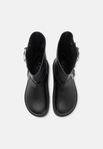 Bottes noires en cuir au niveau de la cheville avec une texture lisse, dotées d'un détail de boucle sur le côté, d'un bout rond et d'un petit talon. La doublure intérieure est sombre.