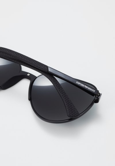 Ochelari de soare negri cu lentile rotunde, brațe texturate negre și detalii metalice. Prezintă lentile întunecate și o ușoară curbură în design.