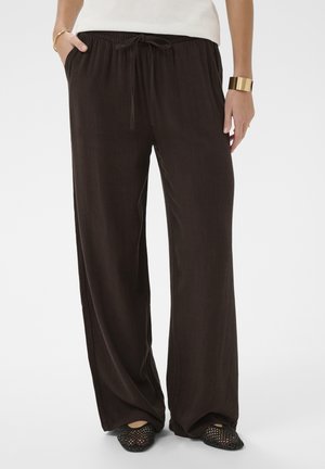 Person iført løstsiddende sorte bukser med snøre og sorte flettede flade sko, stående med den ene hånd i lommen og gyldne armbånd på håndleddene.