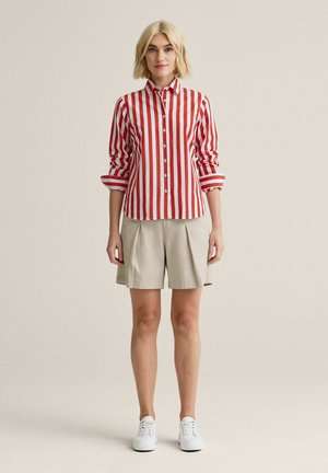 Nainen, jolla on lyhyet vaaleat hiukset, pukeutuneena puna-valkoraidalliseen napitettuun paitaan, beigenvärisiin laskostettuihin shortseihin ja valkoisiin tennareihin.