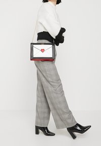 Borsa a tracolla nera, bianca e rossa a forma di busta con un dettaglio a cuore rosso. Presenta una tracolla in catena dorata e una texture in pelle liscia.