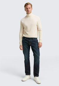 Crème coltrui, donkerblauwe jeans en witte sneakers. De trui heeft een geribbeld patroon; de jeans zijn recht gesneden met contrasterende stiksels.
