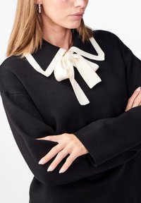 Pull noir avec un col blanc et un détail de nœud papillon. Le tissu semble doux, avec des manches longues et une coupe décontractée.