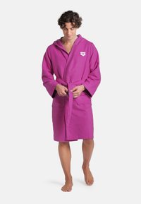 Accappatoio di cotone viola con cappuccio, cintura annodata e tasche frontali. Presenta un logo sul petto e una lunghezza media fino al polpaccio.