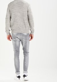 Maglione grigio con motivo a maglia testurizzata, abbinato a jeans grigi chiari con strappi e macchie di vernice, e sneakers bianche con accenti neri.