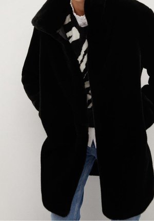 Personne portant un manteau en fausse fourrure noir sur un pull à motifs noir et blanc et un jean bleu, sur un fond uni.