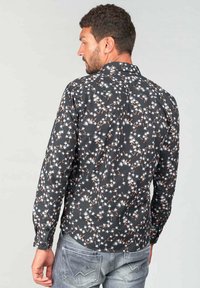 Camisa de manga larga en tela oscura con un patrón floral en blanco y rojo. Presenta un cuello con botones y corte ajustado. Lleva puesta unos jeans claros.