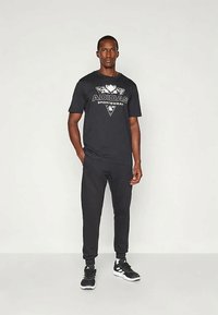 Μαύρο T-shirt Adidas με λευκό λογότυπο, συνδυασμένο με μαύρες φόρμες. Το μοντέλο φοράει μαύρα αθλητικά παπούτσια. Απλός, αθλητικός σχεδιασμός και γραμμή.