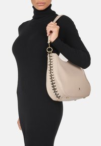 Beżowa skórzana torebka o wyraźnym kształcie, z plecionym detalem po jednej stronie i złotymi akcentami.