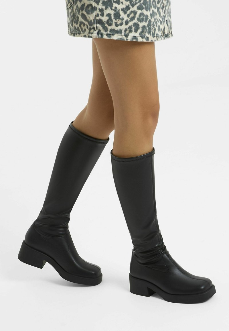Knee-high black boots made of smooth leather with a chunky heel and a slightly slouched design, featuring a sleek and modern silhouette.