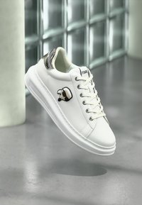 Vit låg sneaker med svart häl detalj, vita snören och en stiliserad tecknad ansikte-logotyp på sidan, svävande över ett grått golv.