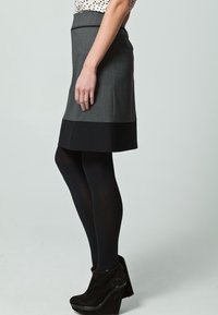 Grauem Rock mit schwarzem Saum, der eine figurbetonte Passform bietet. Das Model trägt schwarze blickdichte Strumpfhosen und schwarze Keilabsatzschuhe.