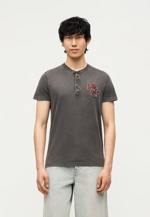 Jeune homme aux cheveux bouclés foncés porte un t-shirt henley noir délavé avec le logo « HCo » sur la poitrine et un jean gris clair, debout devant un fond uni.