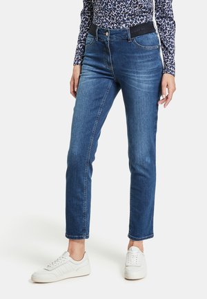 Mujer con vaqueros azules de tiro medio, una camiseta de manga larga con estampado de leopardo y zapatillas blancas, de pie frente a un fondo liso.