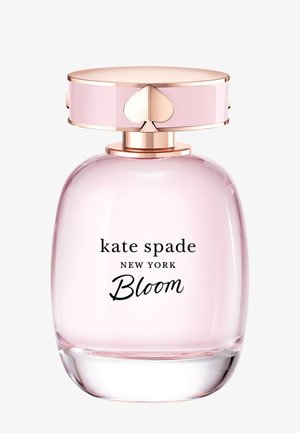 Różowa szklana butelka perfum o zaokrąglonym kształcie z korkiem z złotym akcentem, na którym znajduje się logo w kształcie serca. Etykieta głosi "kate spade NEW YORK Bloom."
