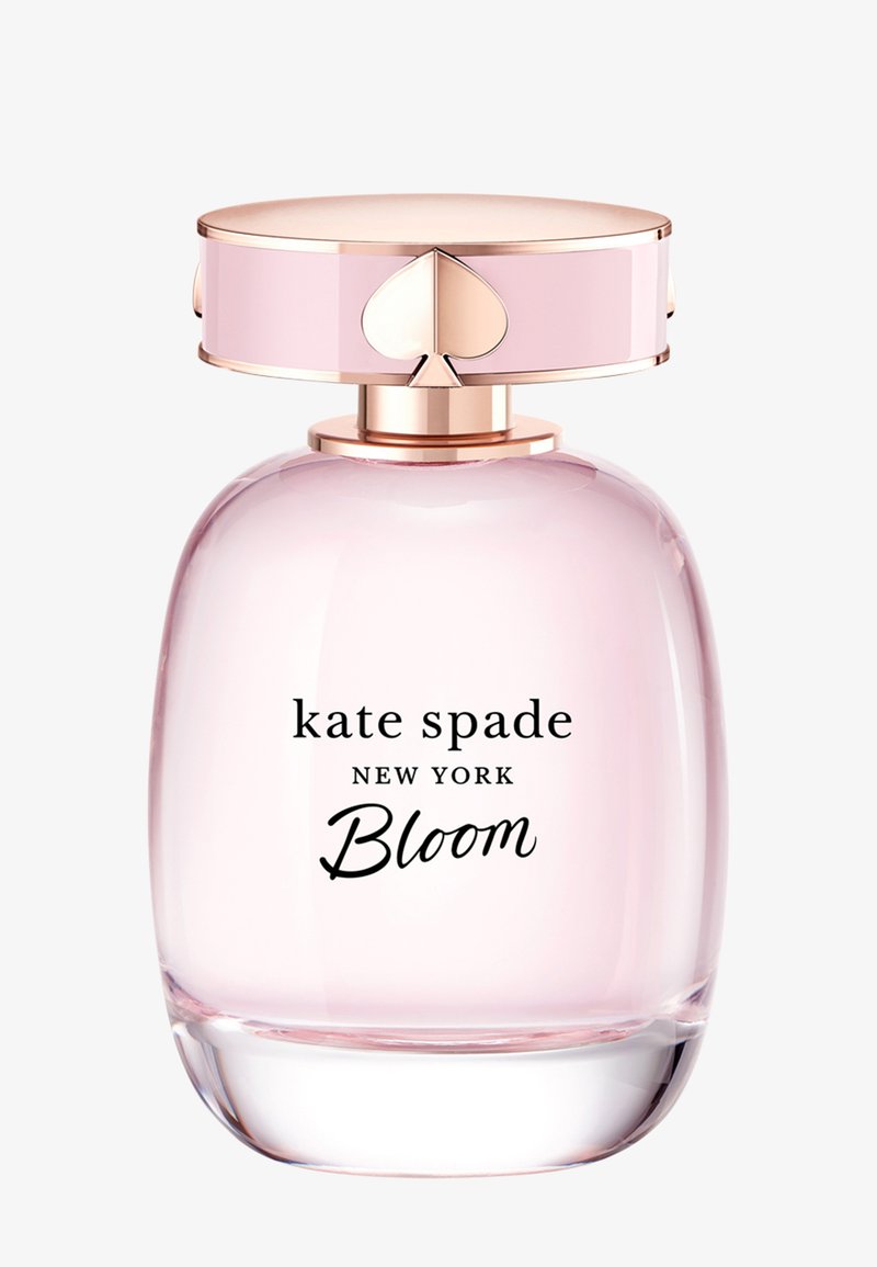 Różowa szklana butelka perfum o zaokrąglonym kształcie z korkiem z złotym akcentem, na którym znajduje się logo w kształcie serca. Etykieta głosi "kate spade NEW YORK Bloom."