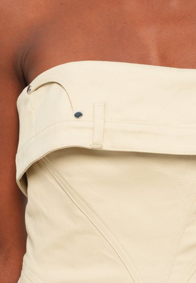 Top beige sem alças com um design estruturado e costuras visíveis. Apresenta detalhes em metal prateado na linha do pescoço e uma textura suave.