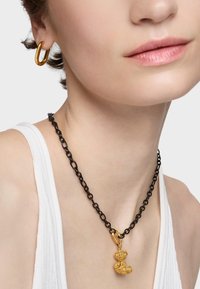Złoty wisiorek w kształcie misia na czarnym naszyjniku, z prostą złotą kolczykiem w kształcie pierścienia. Łańcuszek ma owalne ogniwa, a wisiorek jest fakturowany.