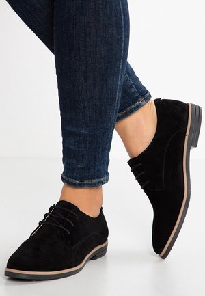 Chaussures à lacets en daim noir avec une semelle en caoutchouc, dotées d'un petit talon et d'un design minimaliste. Portées avec un jean ajusté bleu foncé.
