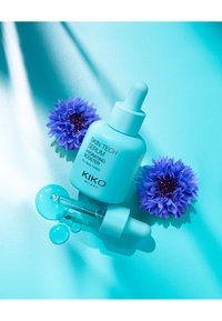 Türkise Flasche mit Pipette, beschriftet "Skin Tech Serum." Begleitet von blauen Blumen und Tropfen auf einer glatten türkisen Oberfläche.