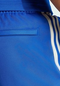 Närbild av blåa träningbyxor med vita och blåa ränder på sidan och en passpojlomme-detalj vid bakre midjan.