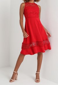 Robe rouge avec un corsage en dentelle et un ourlet inférieur, dotée d'une jupe évasée. Comprend un accent en dentelle texturée près de l'ourlet et portée avec des talons beiges.