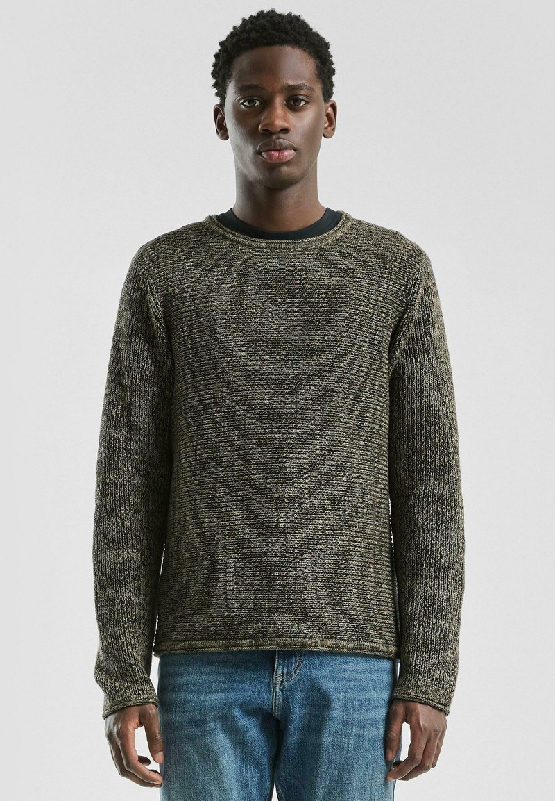 Gebreide trui in donker groen en bruine tinten, met een gestructureerd patroon, ronde halslijn en lange mouwen, gecombineerd met lichtblauwe jeans.