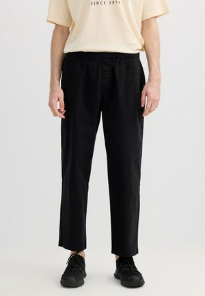 Modelo masculino con pantalones negros de pierna recta con cintura elástica y zapatillas negras, combinado con una camiseta beige.