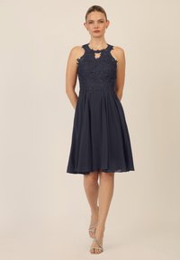 Navyblaues Kleid mit Spitze im Oberteil, Halterneck-Design, fließendem Rock, knielang, mit einem Schlüssel-Ausschnitt an der Vorderseite. Silberne Riemchen-High Heels.