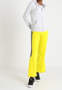 Felpa grigia con zip e tasca frontale, abbinata a pantaloni larghi gialli brillanti con strisce nere laterali e bottoni a pressione, indossata con sneakers nere.
