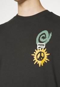 Black t-shirt featuring a green spiral graphic above a yellow sun with a peace symbol, with a "PUMA" logo in white. Cotton material.