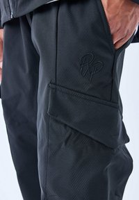 Pantalones cargo negros con material texturizado, que presentan un bolsillo lateral y un logo bordado. Corte ajustado con detalles de costura visibles.