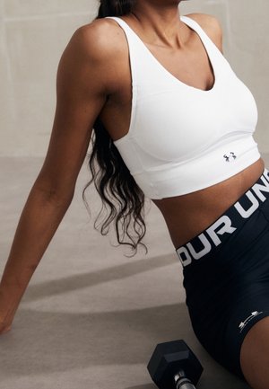 Frau trägt weißen Sport-BH und schwarze Under Armour Shorts, sitzt auf dem Boden neben einer schwarzen Hantel.