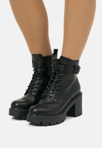 Bottines en cuir noir avec un devant à lacets, fermeture éclair et une sangle ajustable. Talon épais et semelle texturée pour une meilleure adhérence.