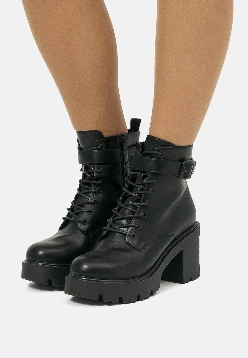 Bottines en cuir noir avec un devant à lacets, fermeture éclair et une sangle ajustable. Talon épais et semelle texturée pour une meilleure adhérence.
