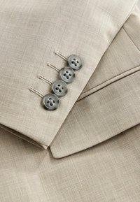 Manche de costume beige avec quatre boutons gris cousus près du poignet et un rabat visible d'une poche assortie.