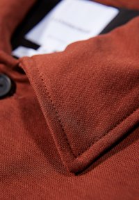 Rostfarbener Stoffkragen mit glatter Textur und sauberer Naht. Enthält ein schwarzes Knopfdetail und ein Pflegeschild, das darunter sichtbar ist.