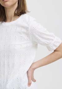 Blusa bianca in cotone con tessuto testurizzato e motivi a occhielli, maniche a sbuffo corte e scollatura rotonda, progettata per favorire la traspirabilità.