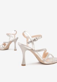 Silberne Riemchensandaletten mit glänzender Oberfläche, schmalen Knöchelriemen und Pfennigabsatz. Offene Zehenpartie mit dekorativen Ausschnitten.