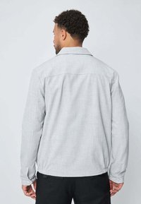 Homme aux cheveux bouclés courts, portant une veste à manches longues gris clair et un pantalon noir, tourné le dos contre un fond uni clair.