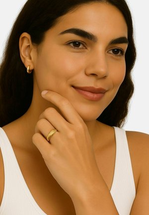 Woman with dark hair wearing a white tank top, gold hoop earrings, and a twisted gold ring, gently touching her chin with a slight smile.
