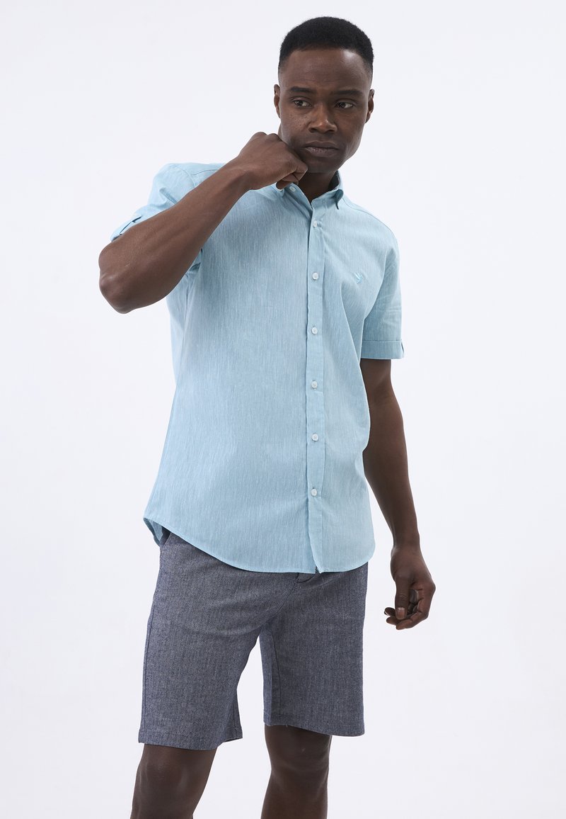 Button-down-Hemdkäppchen in Hellblau, mit kurzen Ärmeln, aus strukturiertem Stoff. Kombiniert mit grauen Shorts, die ein lässiges Sommeroutfit präsentieren.