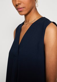 Top sans manches marine avec un décolleté en V profond. Fabriqué en tissu lisse, il présente de simples détails de couture et est accompagné d'un collier en or.