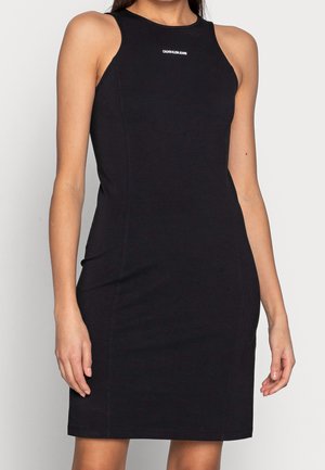 Woman wearing a sleeveless black fitted dress with a high neckline and small "Calvin Klein Jeans" logo on the chest.