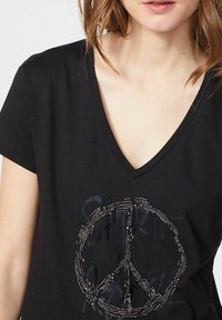 Camiseta de algodón negra con escote en V que presenta un símbolo de paz hecho de tachuelas plateadas, con elementos de texto impresos en un tono más oscuro.