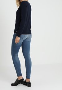 Žena v tmavě modrém tričku s dlouhým rukávem, světle modrých skinny džínách a černých lakovaných botách, stojí na neutrálním světlem pozadí.